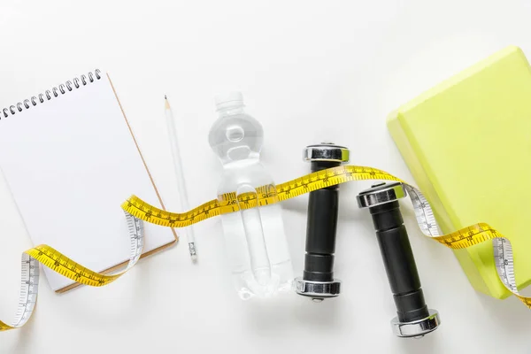 Top view of water in bottle, measuring tape, dumbbells and blank notebook with pencil near box on white background — Stock Photo