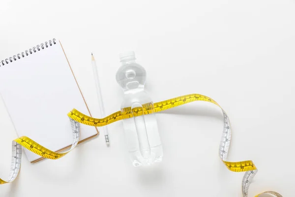 Top view of water in bottle, measuring tape, blank notebook with pencil on white background — Stock Photo