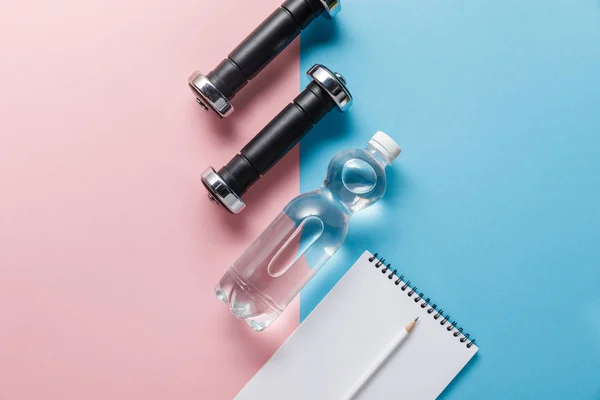 Top view of bottle with water, dumbbells and blank notebook on blue and pink background — Stock Photo