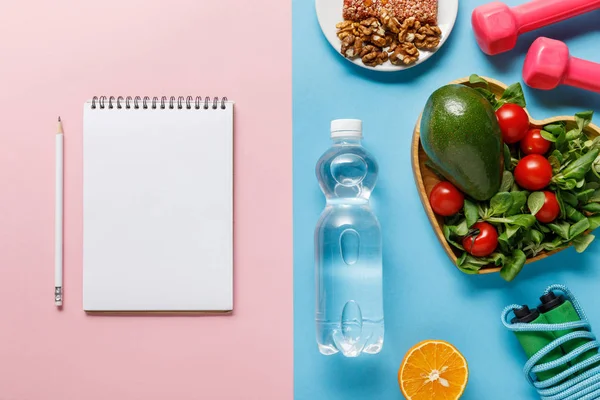 Vista superior de la botella con agua, comida dietética, equipo deportivo y cuaderno en blanco sobre fondo azul y rosa - foto de stock