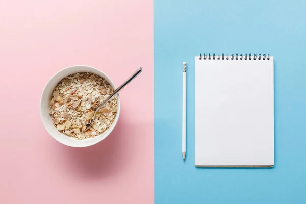 Top view of breakfast cereal in bowl and blank notebook with pencil on blue and pink background — Stock Photo