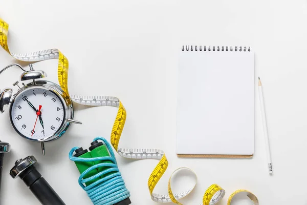 Vista superior del equipo deportivo, cinta métrica, despertador cerca del cuaderno en blanco sobre fondo blanco - foto de stock