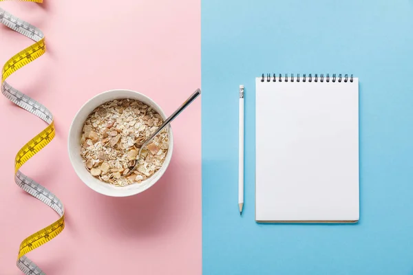 Top view of breakfast cereal in bowl near measuring tape and blank notebook with pencil on blue and pink background — Stock Photo