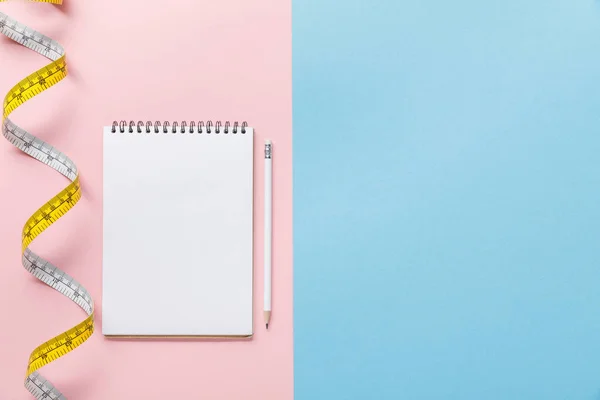 Vista superior de cinta métrica y cuaderno en blanco con lápiz sobre fondo azul y rosa - foto de stock