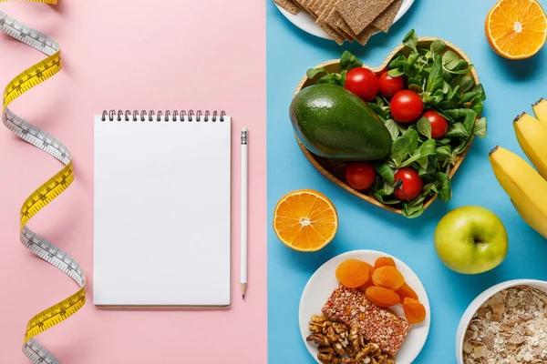 Top view of diet food on blue and empty notebook with pencil and measuring tape on pink background — Stock Photo
