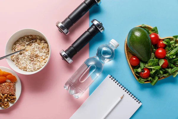 Top view of bottle with water, dumbbells and blank notebook near diet food on blue and pink background — Stock Photo