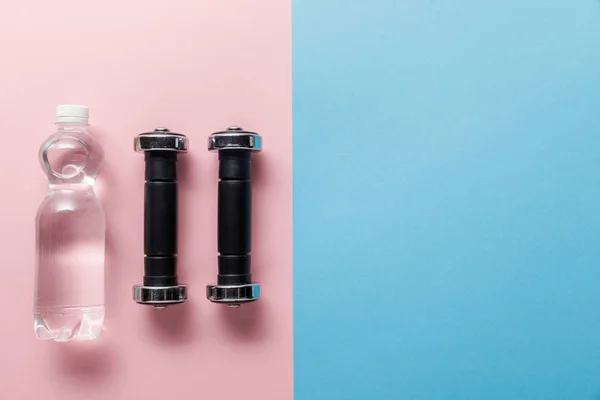 Top view of dumbbells and bottle of water on blue and pink background — Stock Photo