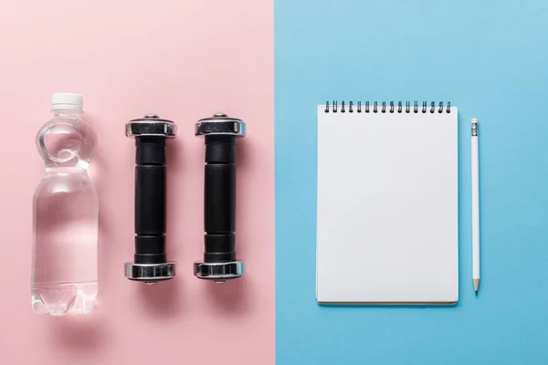 Top view of dumbbells and bottle of water near empty notebook with pencil on blue and pink background — Stock Photo