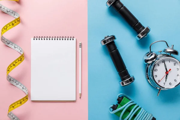 Top view of measuring tape and blank notebook with pencil on blue and alarm clock near skipping rope and dumbbells on pink background — Stock Photo