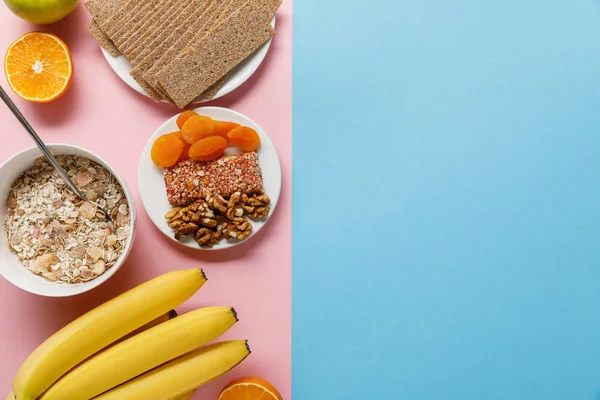 Vista superior de frutas frescas, pan crujiente y cereales de desayuno sobre fondo azul y rosa con espacio para copiar - foto de stock