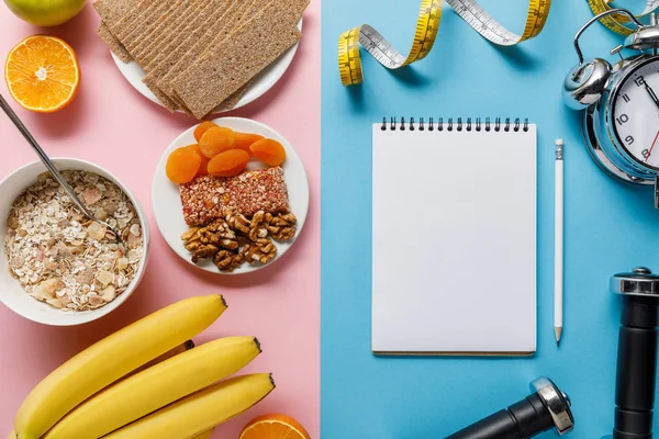 Top view of fresh fruits, crispbread and breakfast cereal on pink and blank notebook, dumbbells and measuring tape on blue background — Stock Photo