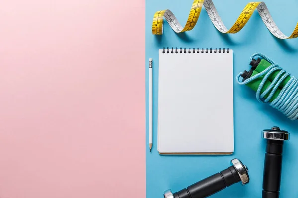 Vista superior de notebook em branco, halteres, pulando corda e fita métrica em fundo azul e rosa — Fotografia de Stock