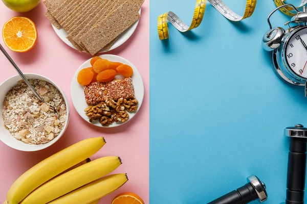 Top view of fresh fruits, crispbread and breakfast cereal on pink and dumbbells and measuring tape on blue background — Stock Photo