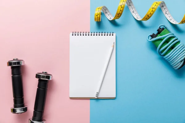 Top view of empty notebook, dumbbells, skipping rope and measuring tape on blue and pink background — Stock Photo