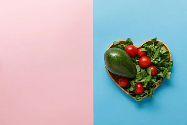 Vista superior de la ensalada de verduras verdes frescas en un tazón en forma de corazón sobre fondo rosa y azul - foto de stock