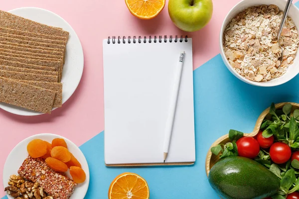 Вид на пустой блокнот и карандаш с вкусной диетической пищей на синем и розовом фоне — стоковое фото