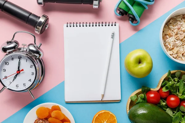Vista superior de cuaderno vacío y lápiz con deliciosa comida dietética y equipo deportivo sobre fondo azul y rosa - foto de stock