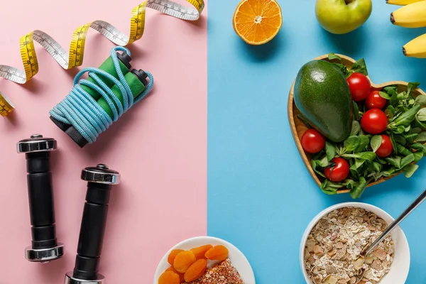 Vista superior de deliciosos alimentos dietéticos e equipamentos esportivos com fita métrica em fundo azul e rosa — Fotografia de Stock