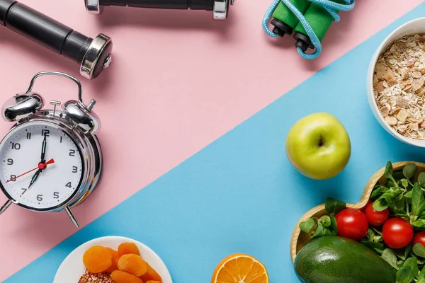 Vista superior de deliciosos alimentos dietéticos e equipamentos esportivos com despertador em fundo azul e rosa — Fotografia de Stock