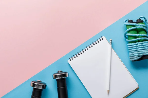 Vista superior de saltar la cuerda, mancuernas y cuaderno en blanco sobre fondo azul y rosa - foto de stock