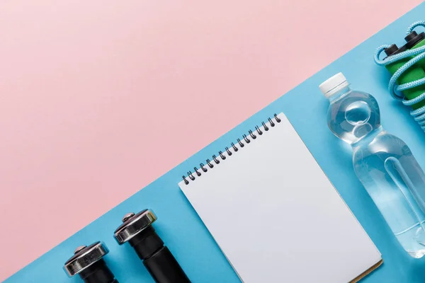 Flat lay with dumbbells, empty notebook, bottle of water and skipping rope on pink and blue background — Stock Photo