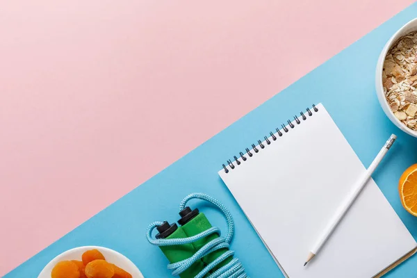 Tendido plano con cuaderno vacío, saltar la cuerda, cereales de desayuno con naranja y albaricoques secos sobre fondo rosa y azul - foto de stock