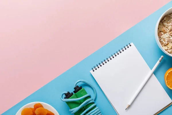 Plat posé avec carnet vide, corde à sauter, céréales pour petit déjeuner avec abricots orange et séchés sur fond rose et bleu — Photo de stock