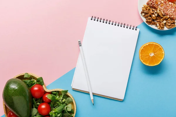 Top view of empty notebook, vegetable salad, orange and nuts on pink and blue background — Stock Photo