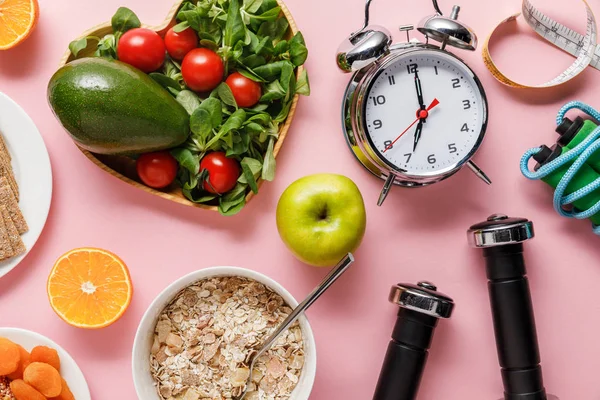 Vista superior de alimentos de dieta fresca, cinta métrica, equipo deportivo y reloj despertador sobre fondo rosa con espacio para copiar - foto de stock