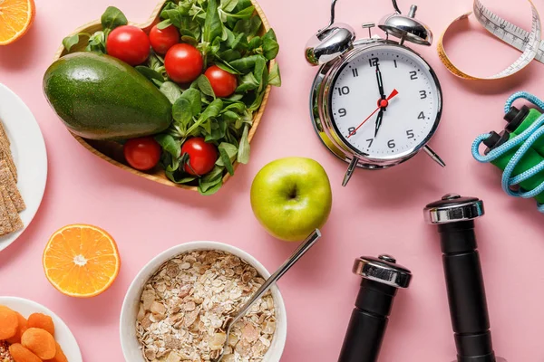 Vue du dessus des aliments frais diététiques, ruban à mesurer, équipement de sport et réveil sur fond rose avec espace de copie — Photo de stock