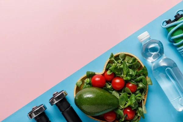 Acostado plano con ensalada de verduras en un tazón en forma de corazón, agua, saltar la cuerda y pesas sobre fondo rosa y azul - foto de stock