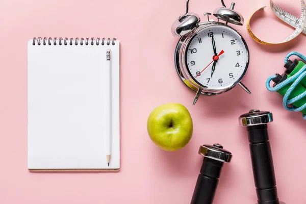 Top view of blank notebook, fresh green apple, dumbbells, measuring tape, skipping rope and alarm clock on pink background with copy space — Stock Photo