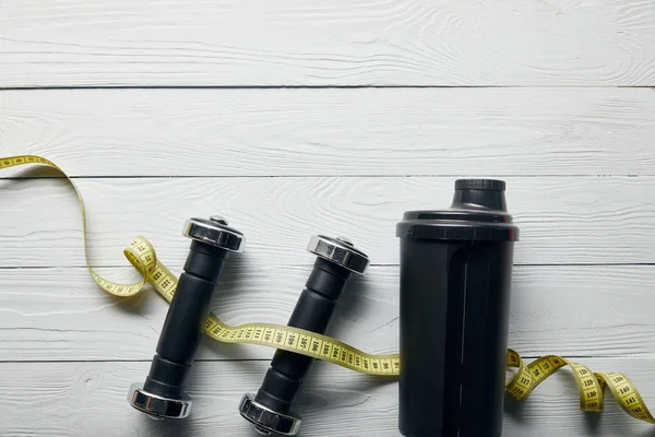 Top view of black shaker cup, measuring tape and dumbbells on wooden white background with copy space — Stock Photo