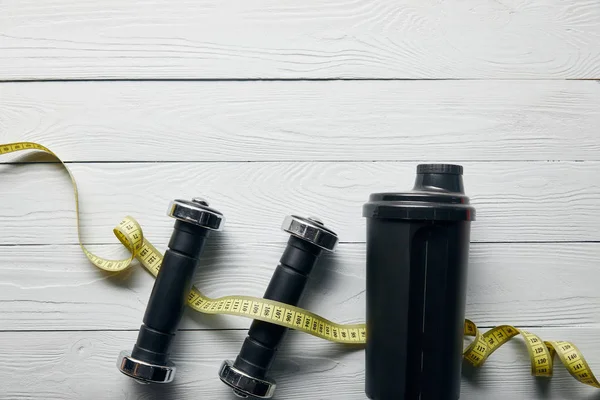 Vista superior de copo shaker preto, fita métrica e halteres em fundo branco de madeira com espaço de cópia — Fotografia de Stock