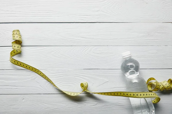 Vue du dessus de la bouteille transparente avec de l'eau et du ruban à mesurer jaune sur fond blanc en bois avec espace de copie — Photo de stock