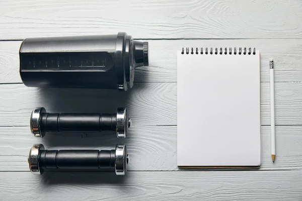 Tendido plano con taza de agitador negro, mancuernas, cuaderno en blanco con espacio para copiar y lápiz sobre fondo blanco de madera - foto de stock