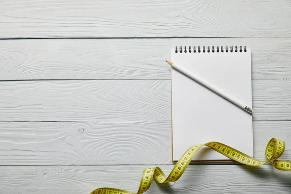 Top view of yellow measuring tape, blank notebook with copy space and pencil on wooden white background — Stock Photo