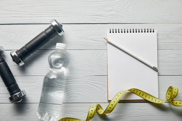 Draufsicht auf Kurzhanteln, Wasser in Flasche, Maßband und Blanko-Notizbuch mit Kopierraum und Bleistift auf weißem Holzgrund — Stockfoto
