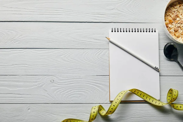 Top view of measuring tape, blank notebook, pencil, spoon and breakfast cereal in bowl on wooden white background — Stock Photo