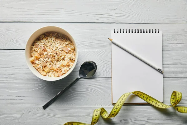 Vista superior de cinta métrica, cuaderno, lápiz, cuchara y cereal para el desayuno en un tazón sobre fondo blanco de madera - foto de stock