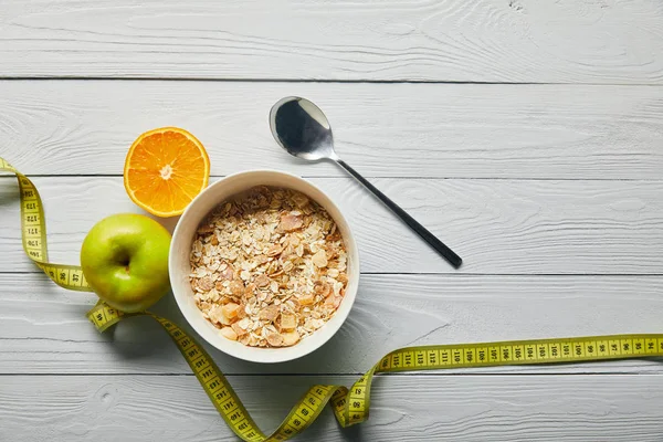Vista superior de cinta métrica, cuchara y cereal de desayuno en tazón cerca de manzana y naranja sobre fondo blanco de madera - foto de stock