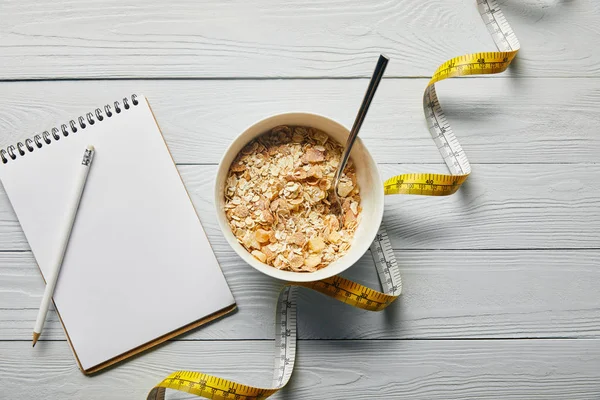 Vista superior de fita métrica, colher e cereal café da manhã em tigela perto de notebook e lápis sobre fundo branco de madeira — Fotografia de Stock