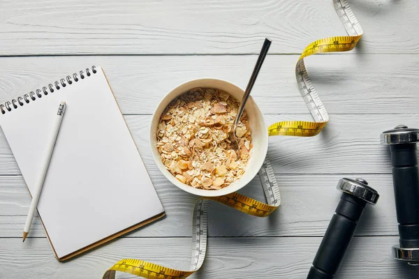 Top view of measuring tape, spoon and breakfast cereal in bowl near notebook, dumbbells and pencil on wooden white background — Stock Photo