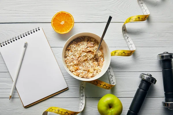 Vista superior de cinta métrica, cuchara y cereal de desayuno en tazón cerca de manzana, naranja, cuaderno, mancuernas y lápiz sobre fondo blanco de madera - foto de stock