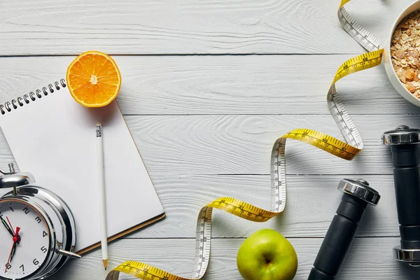 Vista superior de cinta métrica, cereal de desayuno en tazón cerca de manzana, naranja, cuaderno, mancuernas, reloj despertador y lápiz sobre fondo blanco de madera con espacio para copiar - foto de stock