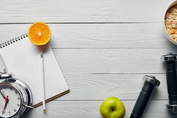 Vue du dessus des céréales pour petit déjeuner dans un bol près de pomme, orange, ordinateur portable, haltères, réveil et crayon sur fond blanc en bois avec espace de copie — Photo de stock