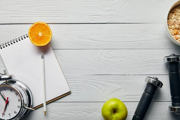 Top view of breakfast cereal in bowl near apple, orange, notebook, dumbbells, alarm clock and pencil on wooden white background with copy space — Stock Photo