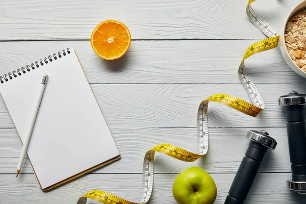 Vista superior de cinta métrica, cereal de desayuno en tazón cerca de manzana, naranja, cuaderno, mancuernas y lápiz sobre fondo blanco de madera con espacio para copiar - foto de stock