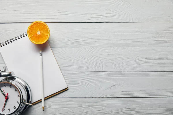 Top view of orange half, notebook and pencil near alarm clock on wooden white background with copy space — Stock Photo