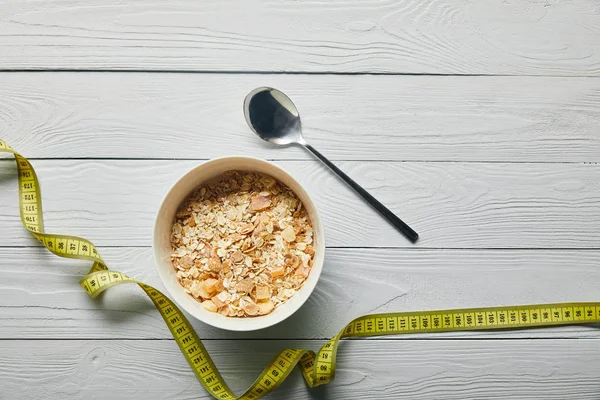 Vista dall'alto del nastro di misurazione, cereali per la colazione in ciotola vicino cucchiaio su sfondo bianco in legno con spazio copia — Foto stock